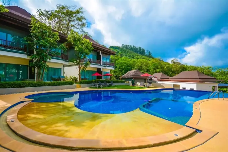 Piscine extérieure avec bâtiments et végétation luxuriante en arrière-plan.
