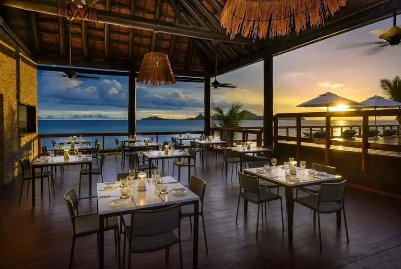 Une salle à manger extérieure avec des tables dressées face à la mer au coucher du soleil.