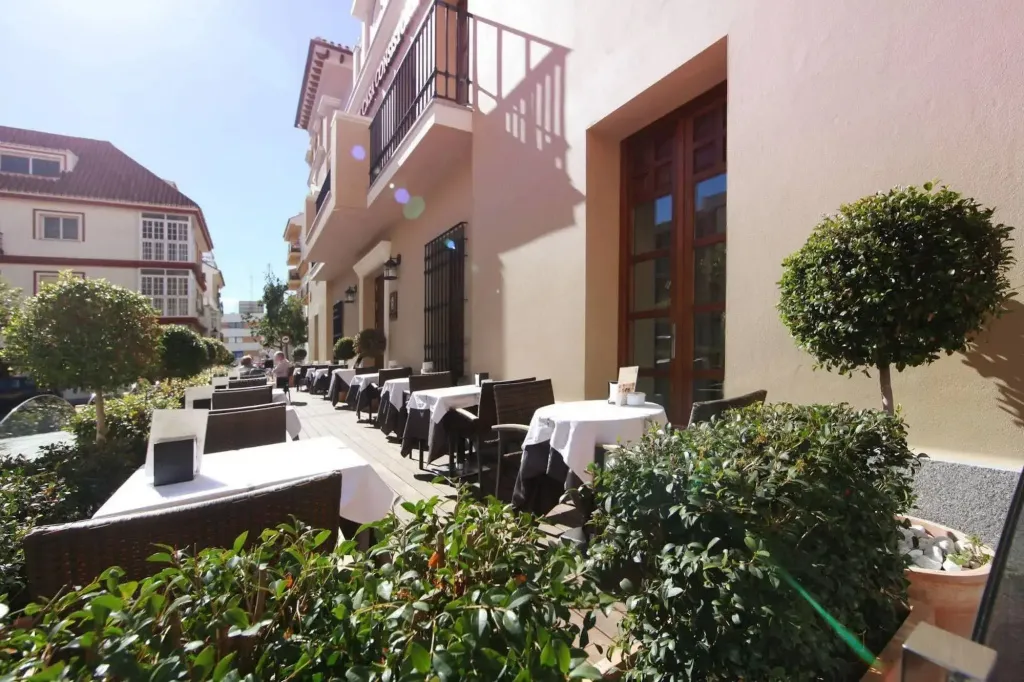 Terrasse ombragée avec tables nappées et chaises en osier devant un bâtiment beige.