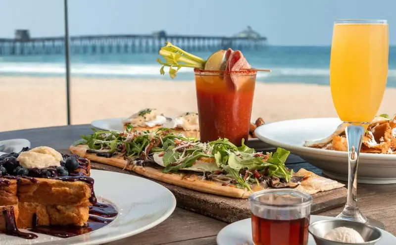 Une table est dressée avec divers plats et boissons, face à une plage et un ponton.