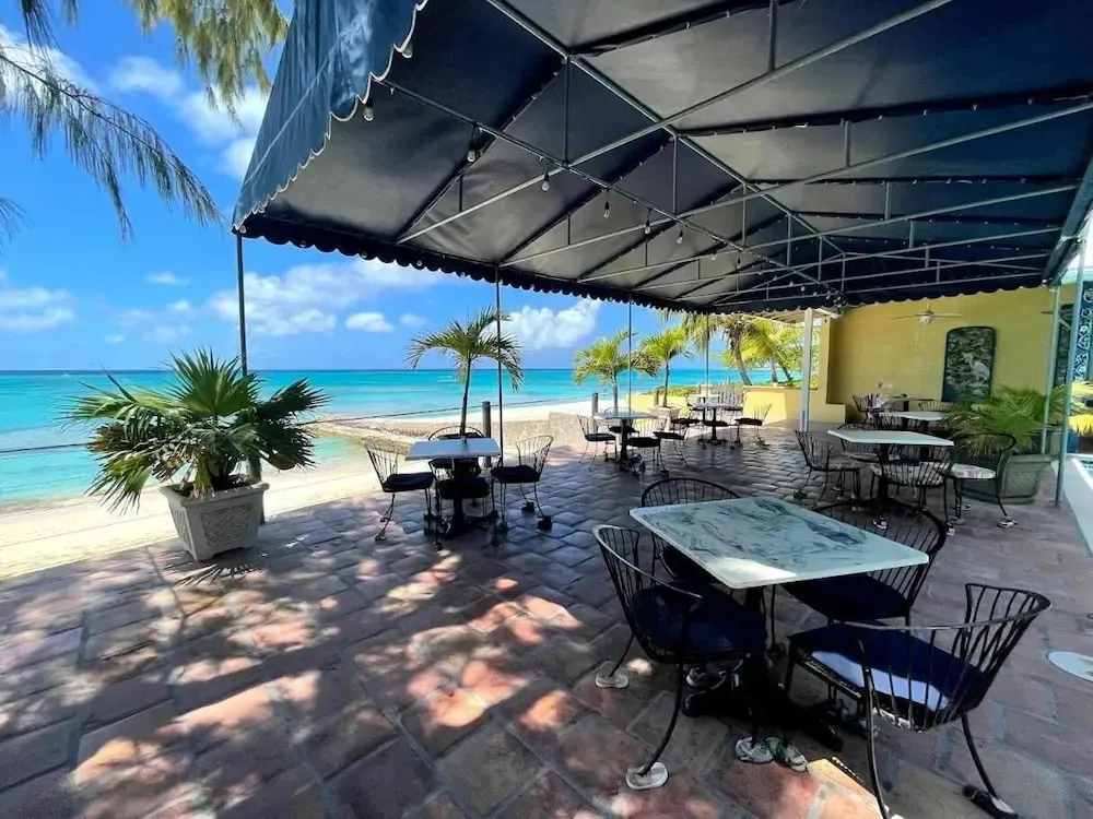 Terrasse ombragée avec tables et chaises donnant sur une plage de sable blanc et une mer turquoise.