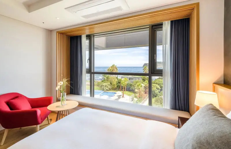 Une chambre lumineuse avec un lit et un fauteuil face à une grande fenêtre donnant sur la mer et une piscine.