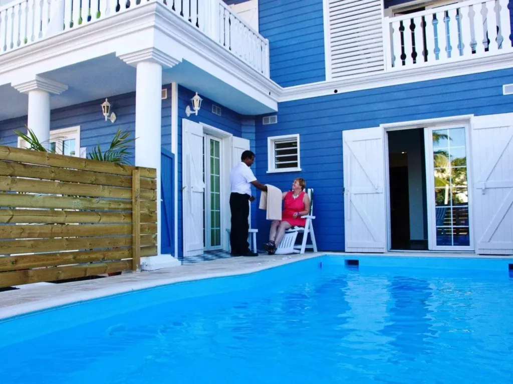 Une piscine turquoise au premier plan, avec un bâtiment bleu et blanc et une personne assise sur une chaise.