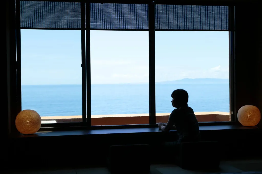 Une personne est assise devant une grande fenêtre donnant sur la mer.