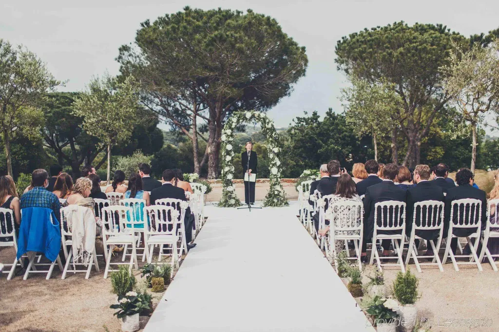 Une cérémonie de mariage se déroule en extérieur, avec des invités assis sur des chaises blanches.