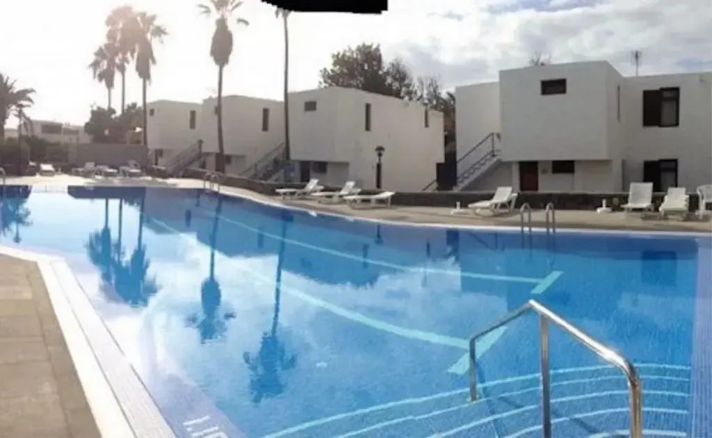 Une piscine bleue bordée de transats blancs, avec des bâtiments blancs en arrière-plan.