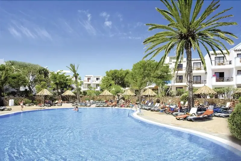 Une piscine ensoleillée bordée de transats et de parasols, avec des bâtiments blancs en arrière-plan.