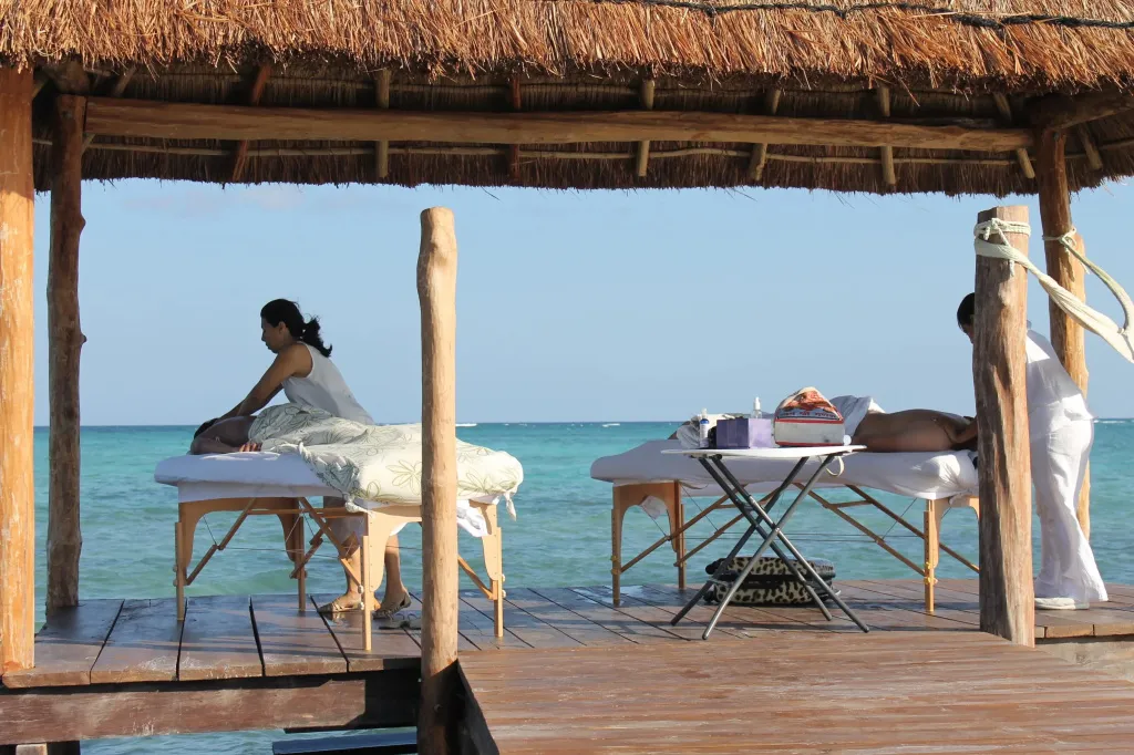Deux personnes allongées sur des tables de massage en bord de mer sous une structure en chaume.
