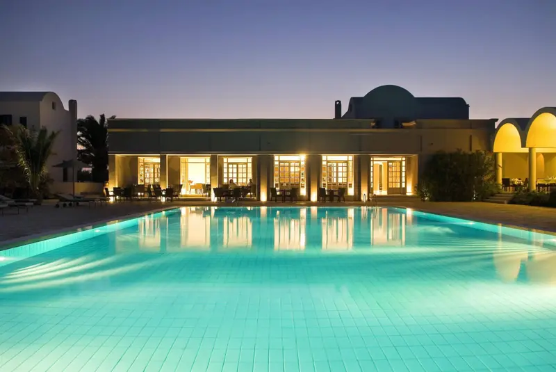 Une piscine illuminée reflète les bâtiments d'un complexe hôtelier au crépuscule.