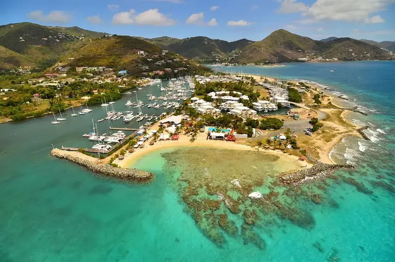 Vue aérienne d'une plage bordée de yachts et entourée de collines verdoyantes.