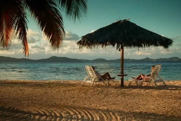 Deux transats invitent à la détente sur une plage de sable fin, ombragée par un parasol en feuilles de palmier.