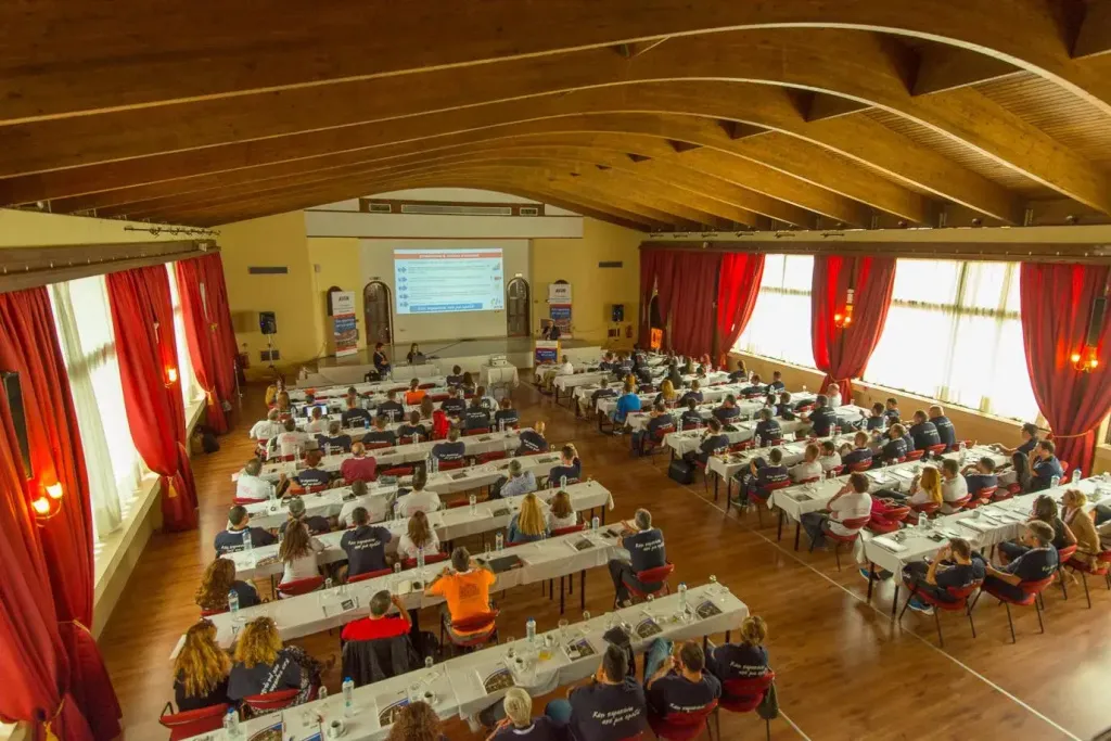 Une grande salle lumineuse accueille une conférence avec de nombreuses personnes assises à des tables.