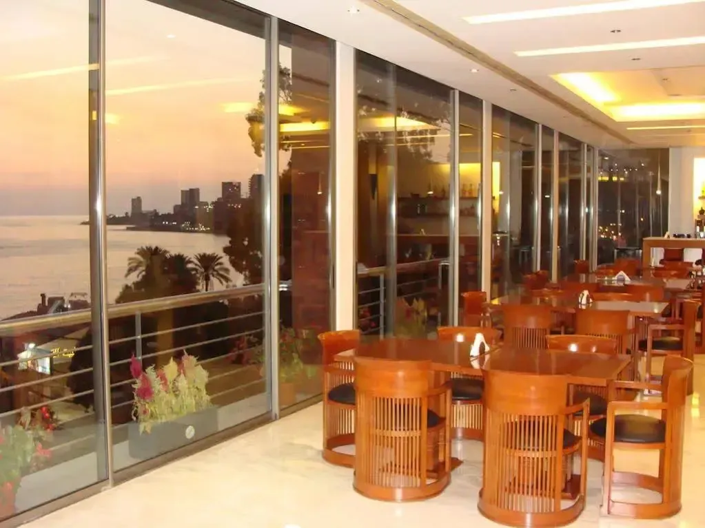 Une salle lumineuse avec des tables et des chaises offre une vue sur la mer et la ville au coucher du soleil.
