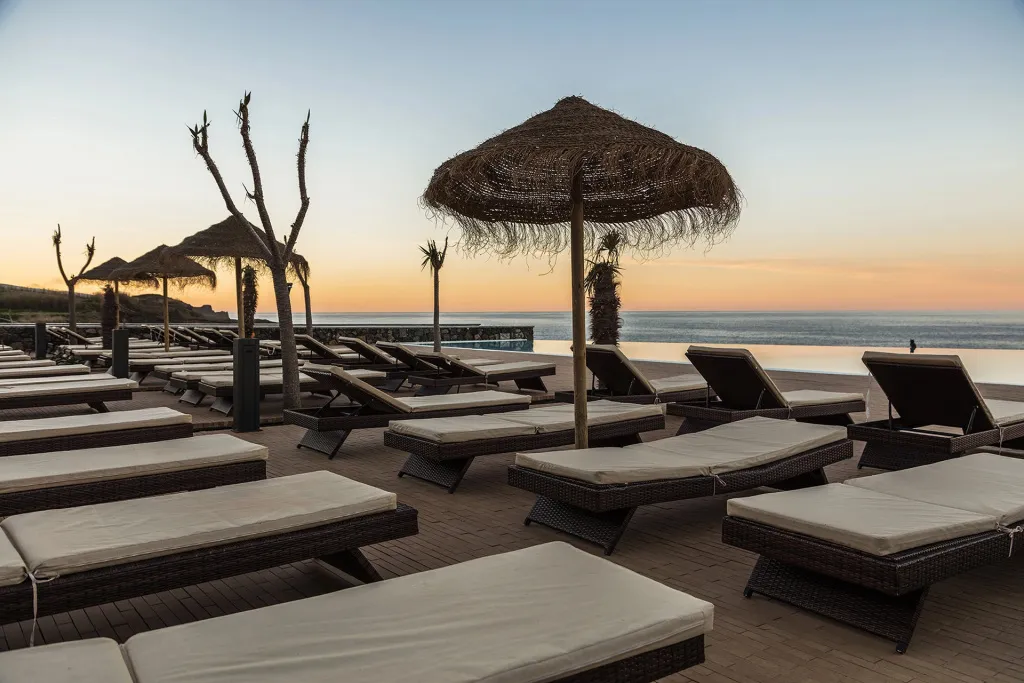 Transats et parasols en chaume bordent une piscine à débordement face à la mer au coucher du soleil.