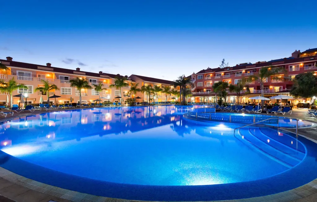 Une piscine illuminée au crépuscule devant un bâtiment à plusieurs étages.