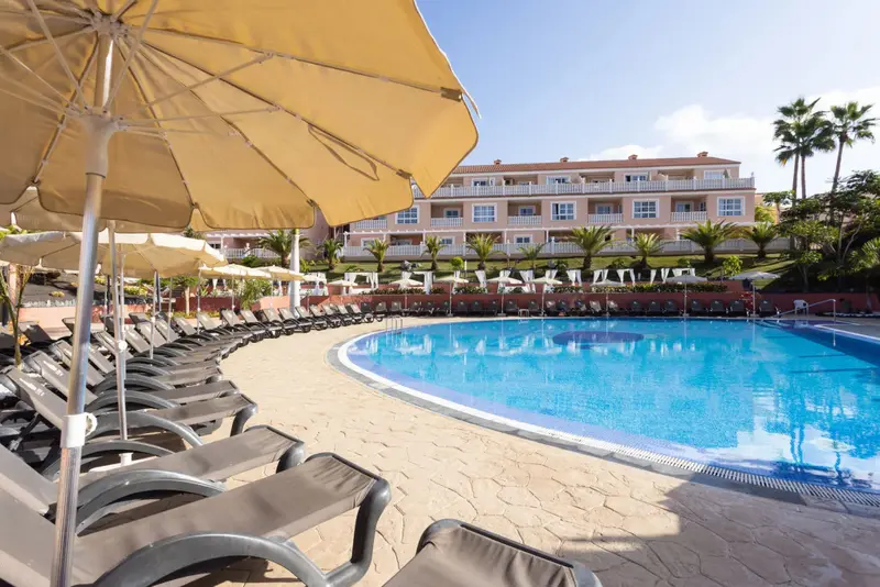 Transats et parasols bordent une piscine turquoise devant un bâtiment beige.