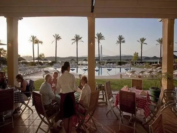 Une terrasse ombragée offre une vue sur une piscine et des palmiers au coucher du soleil.