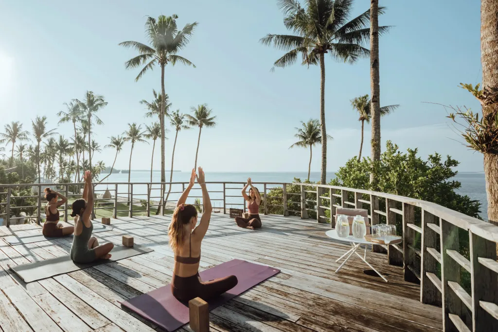Des personnes pratiquent le yoga sur une terrasse en bois avec vue sur la mer et les palmiers.