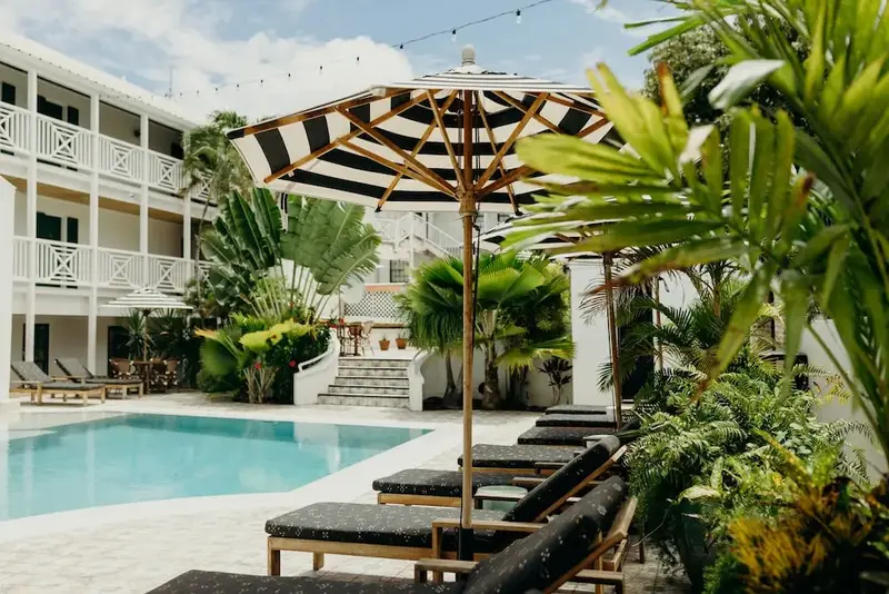 Transats noirs et parasols rayés ombragent une piscine turquoise dans un jardin tropical.