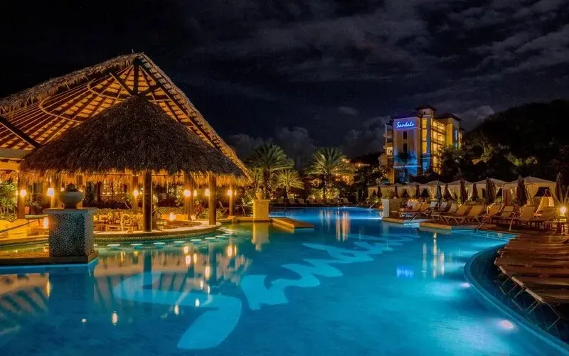 Une piscine illuminée avec un bar en chaume et des transats.