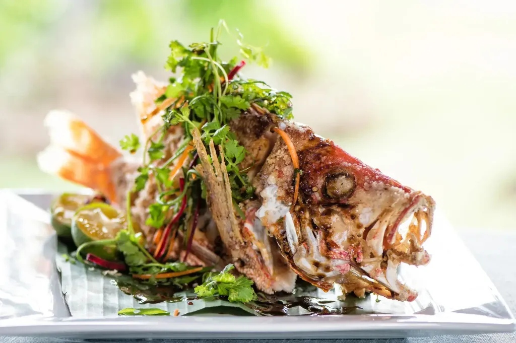 Un poisson grillé est présenté sur une assiette blanche, décoré de verdure et d'agrumes.
