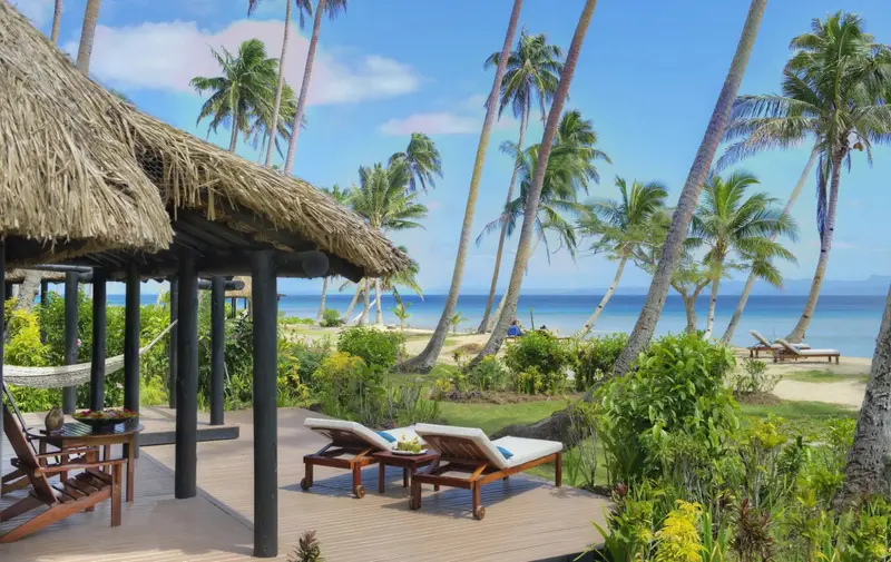Terrasse en bois avec transats et hamac donnant sur une plage de sable fin bordée de palmiers.