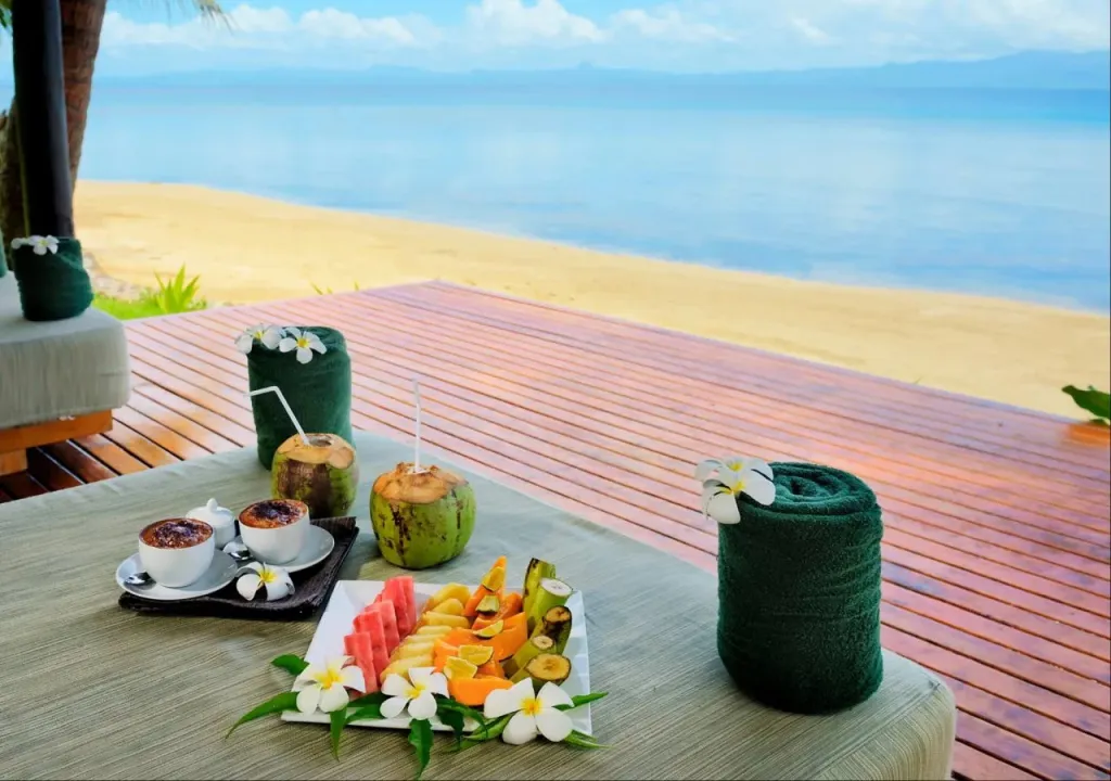 Une table basse est dressée avec des fruits frais et des boissons sur une terrasse en bois face à la mer.