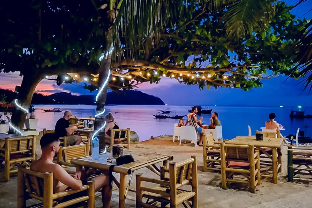 Des convives sont assis à des tables en bois sur une terrasse face à la mer au coucher du soleil.