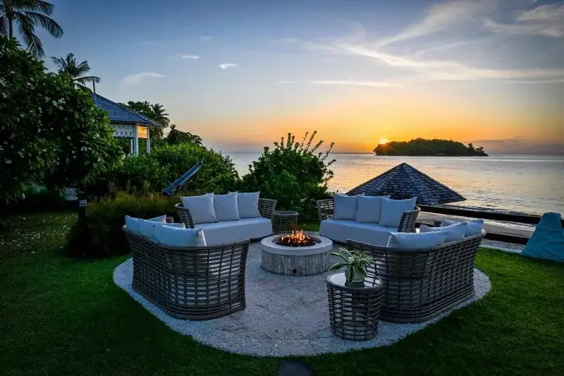 Espace extérieur aménagé avec des fauteuils et un foyer, face à la mer au coucher du soleil.