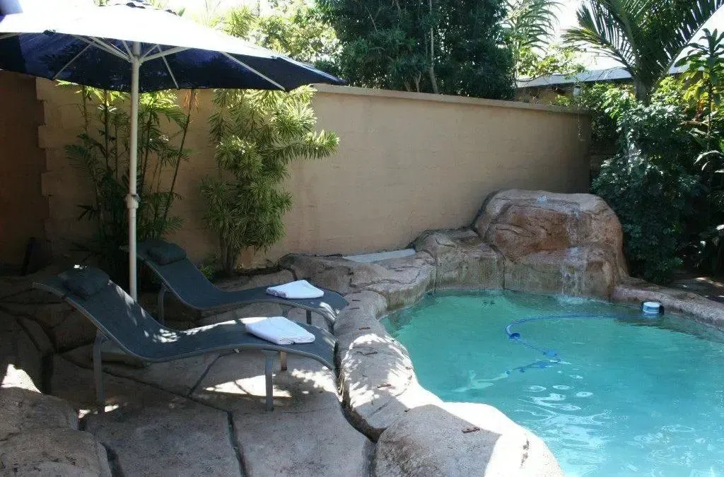 Transats et parasol ombragent une piscine rocheuse dans un jardin luxuriant.