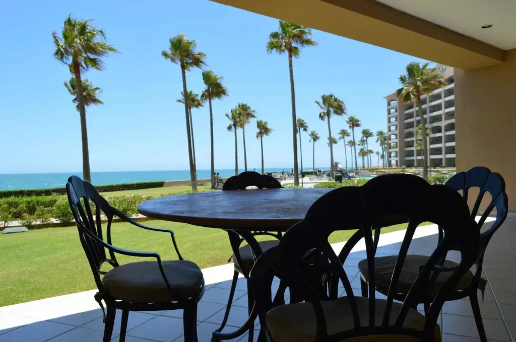 Une table et des chaises invitent à la détente sur une terrasse ensoleillée avec vue sur la mer et les palmiers.