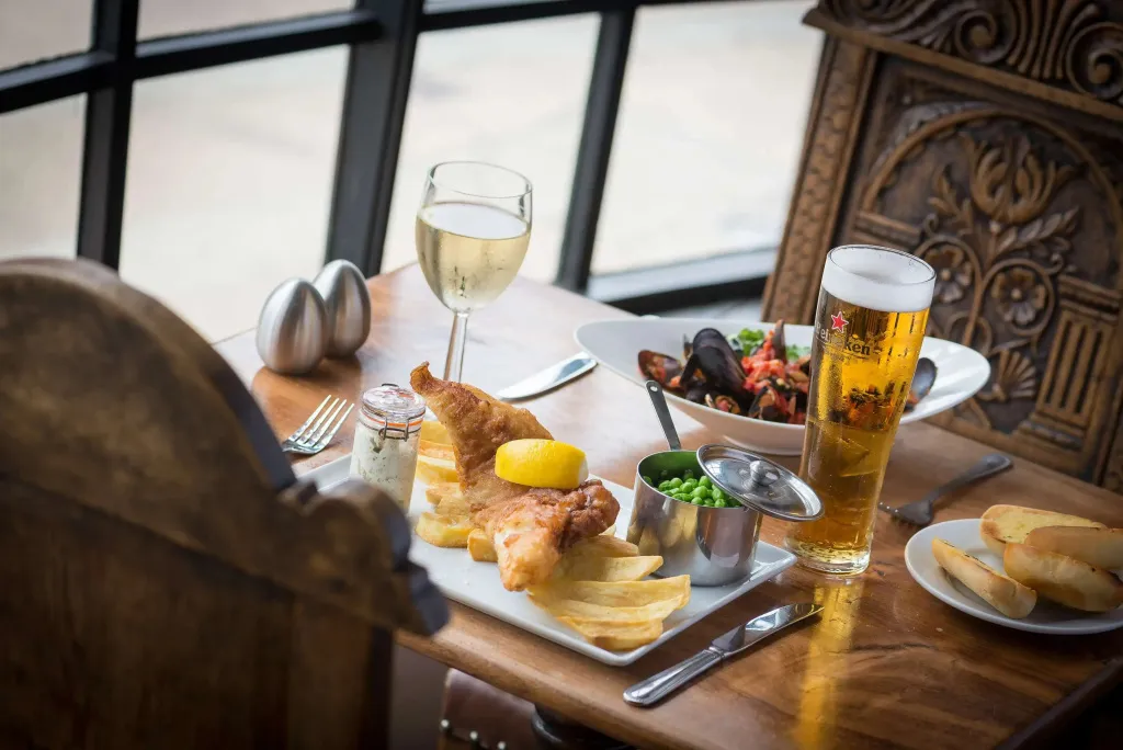 Une table en bois présente des plats de poisson et frites accompagnés de boissons.