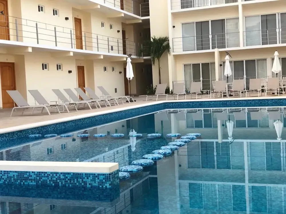 Une piscine rectangulaire bordée de transats et de parasols, devant un bâtiment beige à balcons.