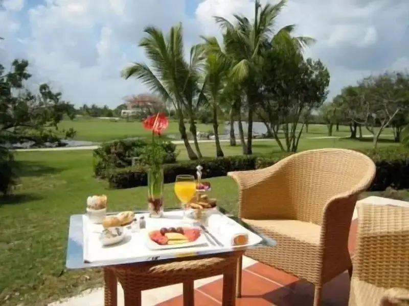 Une table basse en osier présente un petit-déjeuner avec des fruits, des viennoiseries et du jus d'orange sur une terrasse.