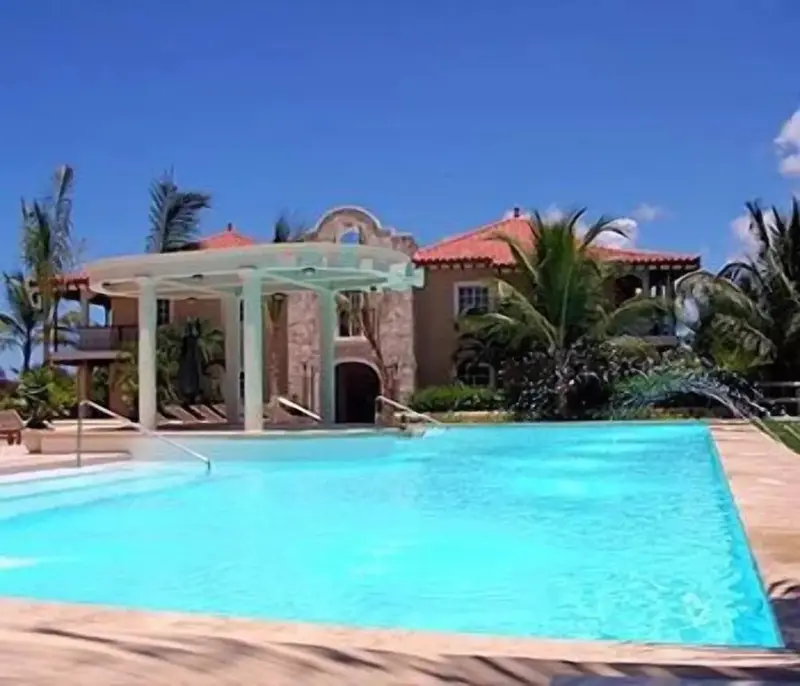 Une piscine rectangulaire turquoise au premier plan, avec un bâtiment aux toits rouges en arrière-plan.