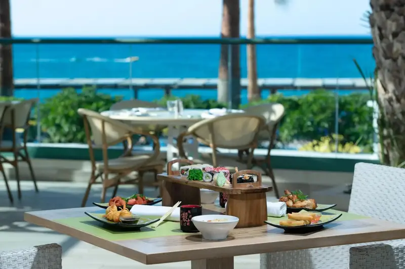 Une table est dressée avec des plats de sushi et des boissons, devant un décor tropical et une vue sur la mer.
