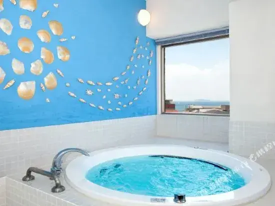 Une baignoire d'angle blanche se trouve dans une salle de bain carrelée, avec une vue sur la mer par la fenêtre.