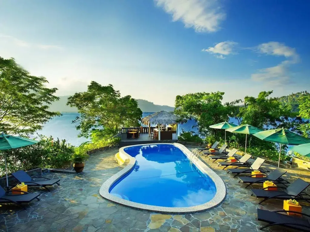 Une piscine bleue bordée de transats et de parasols offre une vue sur la mer.