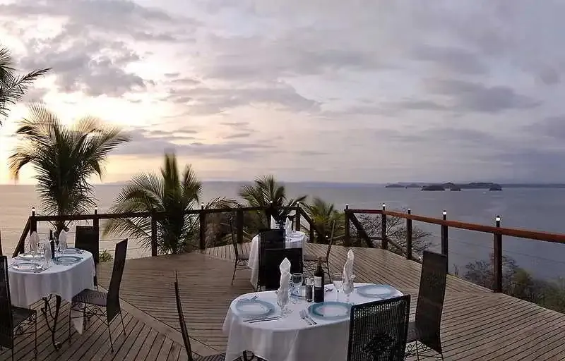Tables dressées sur une terrasse en bois avec vue sur la mer au coucher du soleil.