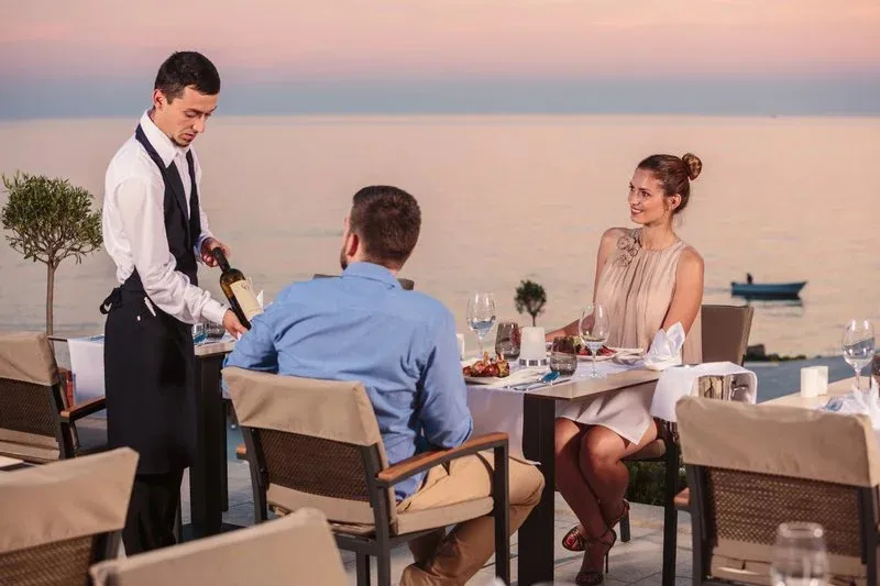 Un serveur verse du vin à des clients attablés en terrasse face à la mer.