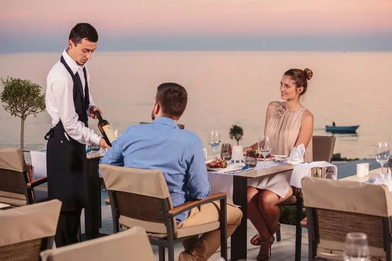 Un serveur verse du vin à des clients attablés en terrasse face à la mer.