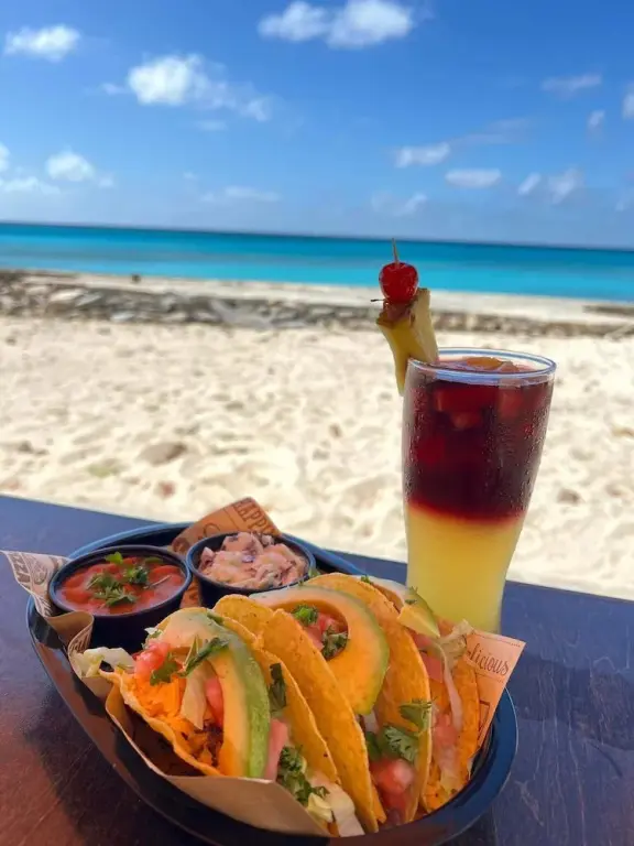 Assiette de tacos et cocktail exotique sur une table en bois face à une plage de sable blanc.