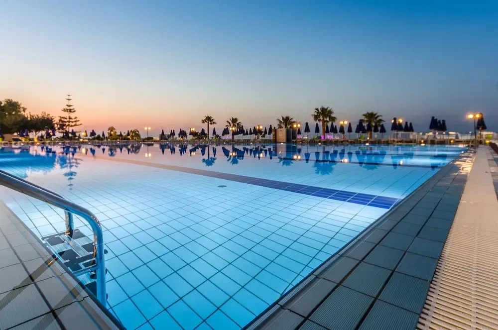 Une piscine rectangulaire illuminée au crépuscule, avec des transats et des parasols en arrière-plan.