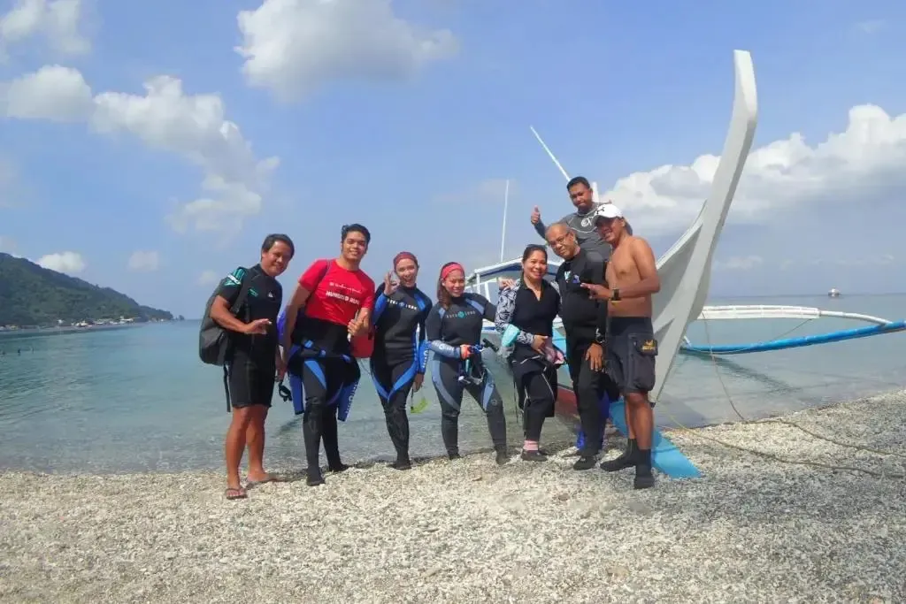 Groupe de personnes souriantes sur une plage de galets, devant un bateau à voile.