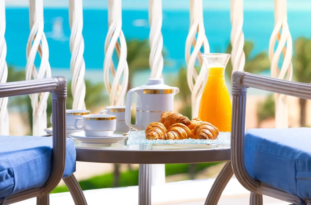 Une table ronde en verre présente un service à thé et des viennoiseries sur une terrasse.