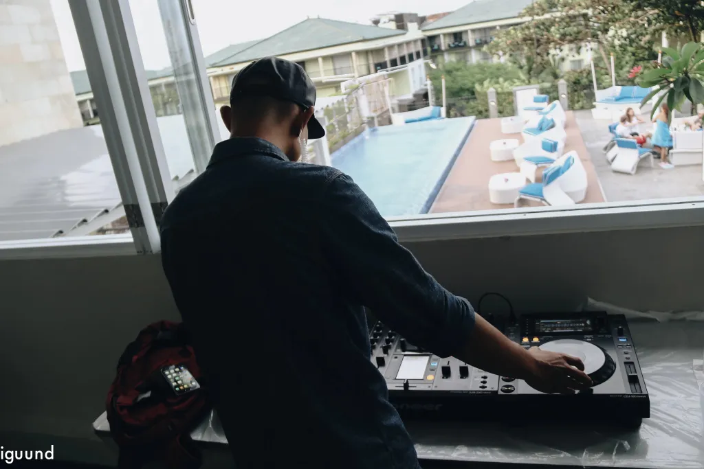 Un homme manipule une platine vinyle près d'une fenêtre donnant sur une piscine et des transats.