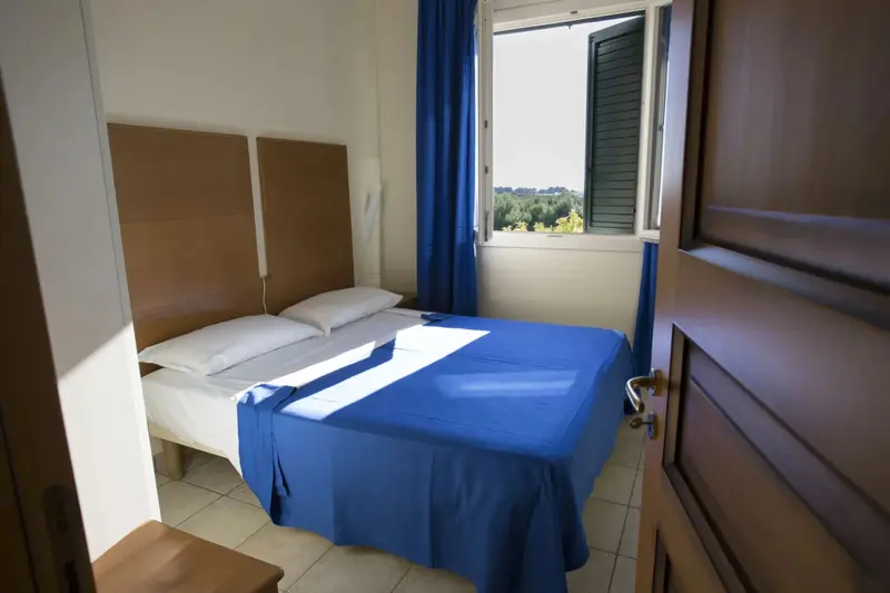 Une chambre lumineuse avec un lit recouvert d'une couverture bleue et une fenêtre ouverte sur un paysage verdoyant.