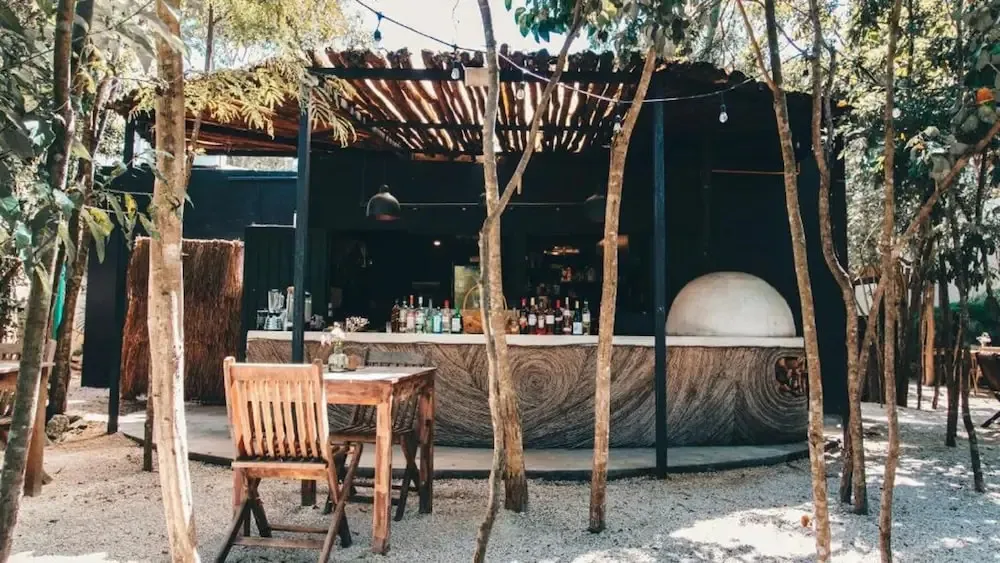 Un bar extérieur ombragé propose des boissons et des places assises sur une terrasse en gravier.
