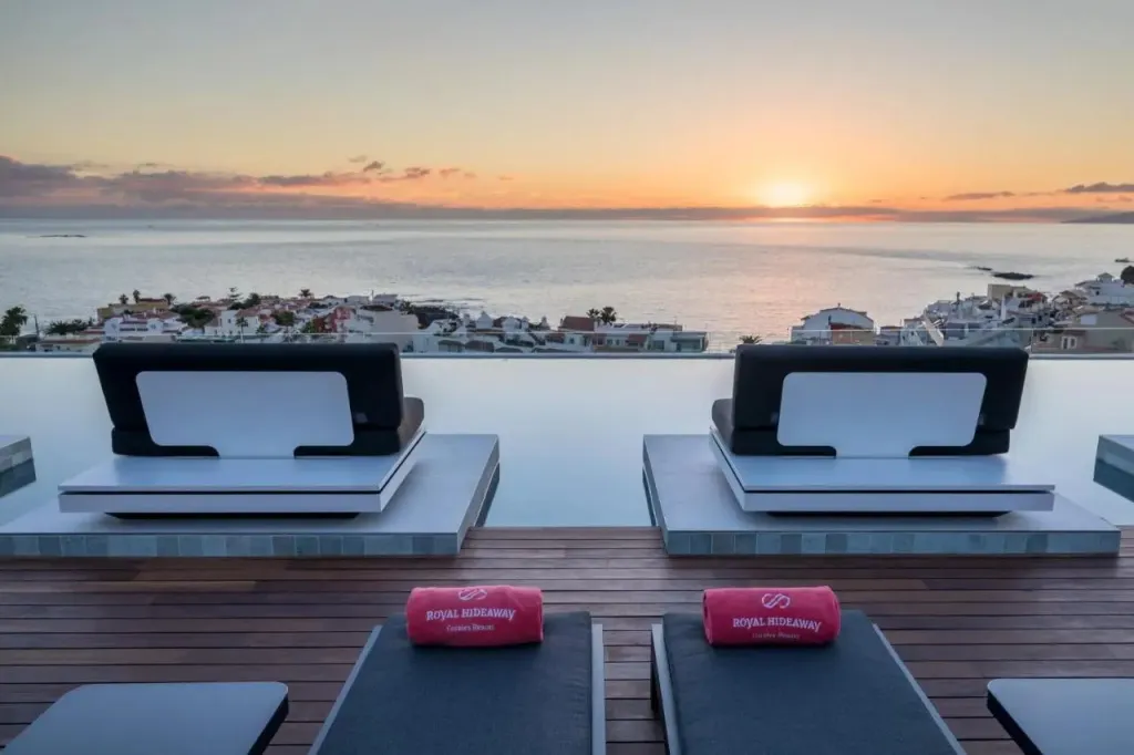 Deux transats avec des serviettes enroulées sont posés au bord d'une piscine à débordement face à la mer au coucher du soleil.