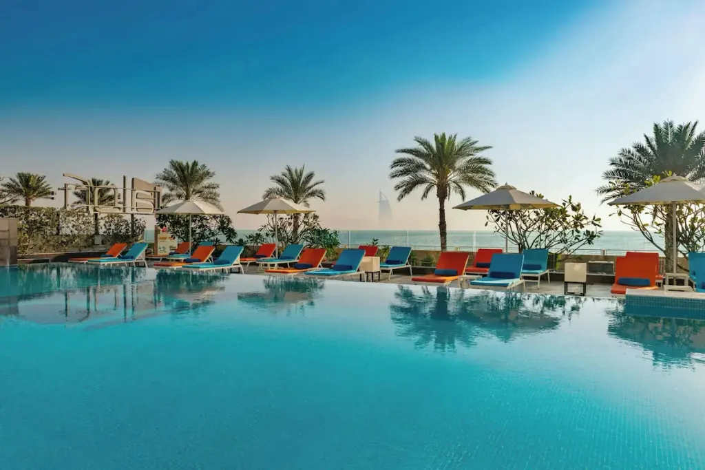 Transats colorés alignés au bord d'une piscine turquoise avec vue sur la mer et des palmiers.